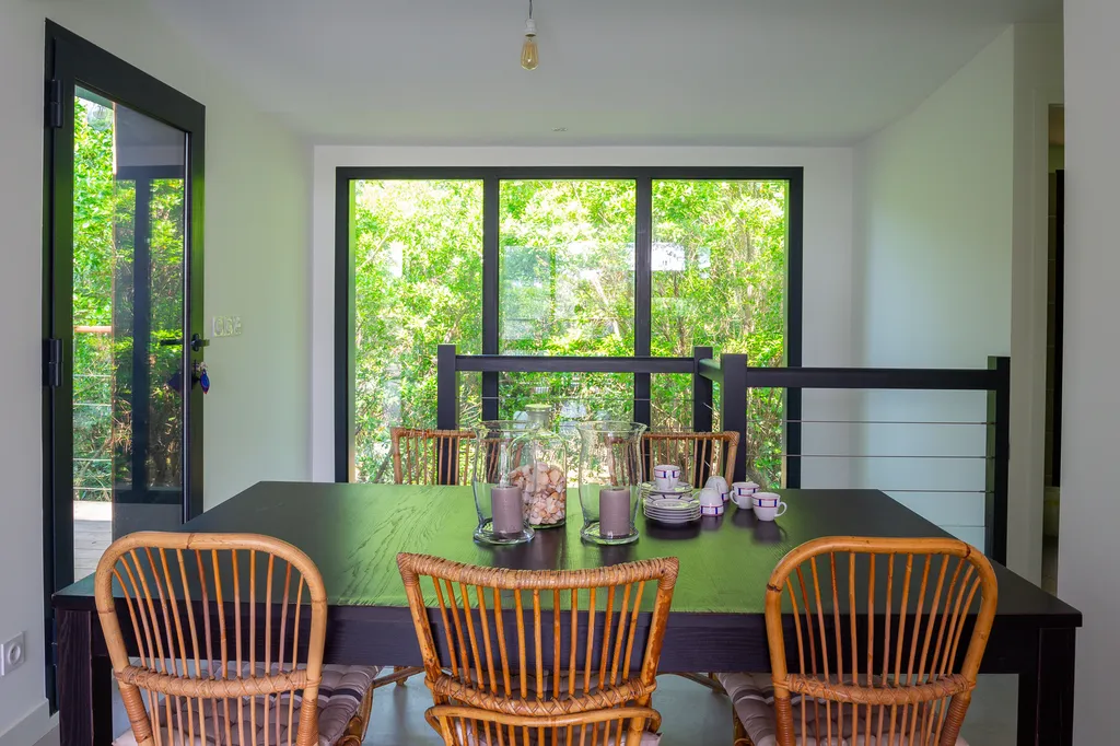 Salle à manger réaménagée autour d’un cadre sur la nature  AKIKO ARCHITECTURE — Piraillan, Cap-Ferret — rénovation intérieure La salle à manger, redessinée sous un ton sable et bois blond, profite d’une ouverture généreuse cadrant la canopée. Le mobilier 
