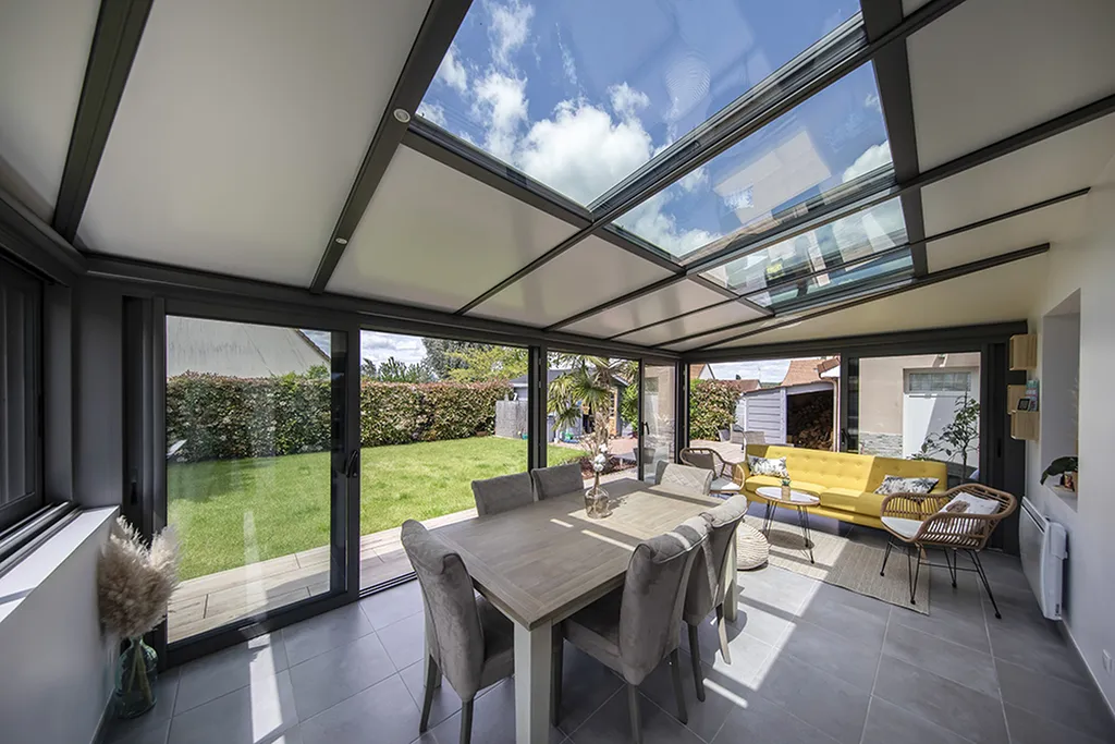 Véranda en aluminium anthracite à toiture mi-vitrée abritant une salle à manger et un coin salon avec canapé jaune à Fréjus et Roquebrune-sur-Argens