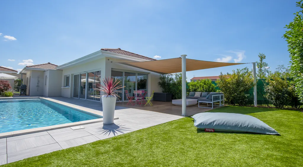 Aménagement paysager avec terrasse bois, gazon synthétique et voiles d’ombrage autour d’une piscine à Genas (69740)