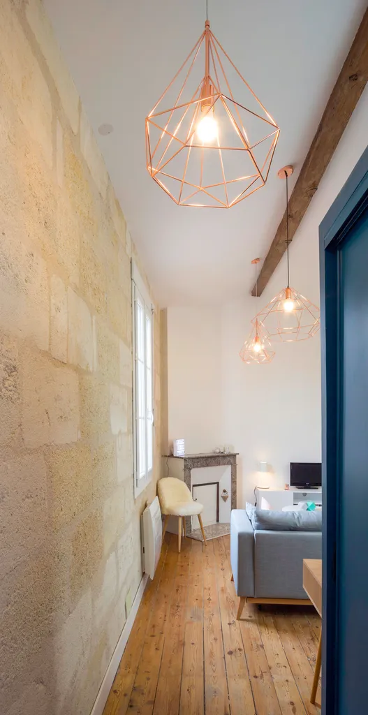 AKIKO ARCHITECTURE – Bordeaux Chartrons — Rénovation & réagencement sous combles Ce couloir, autrefois sombre, a été réouvert et clarifié, laissant apparaître la pierre blonde et un alignement de suspensions cuivrées. La perspective mène naturellement ver